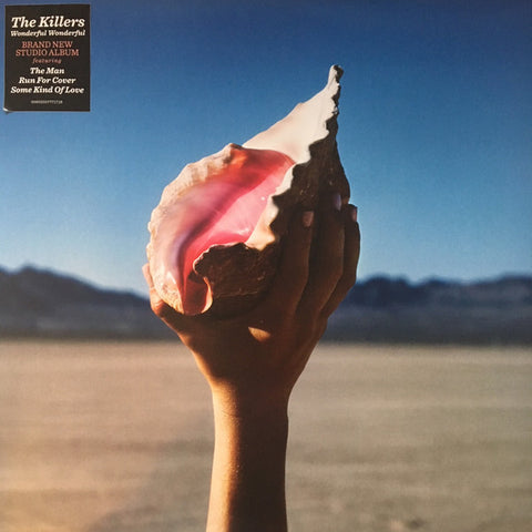 The image shows the cover of the album 'Wonderful Wonderful' by The Killers. It features a hand holding a seashell against a backdrop of a dry, arid landscape under a clear sky.
